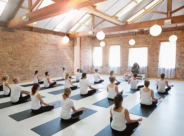 A group of people in a spacious studio practicing yoga seated on mats. They wear white tops and black bottoms, facing a brick wall with windows and round hanging lights. The room has a wooden beam ceiling and a serene atmosphere.