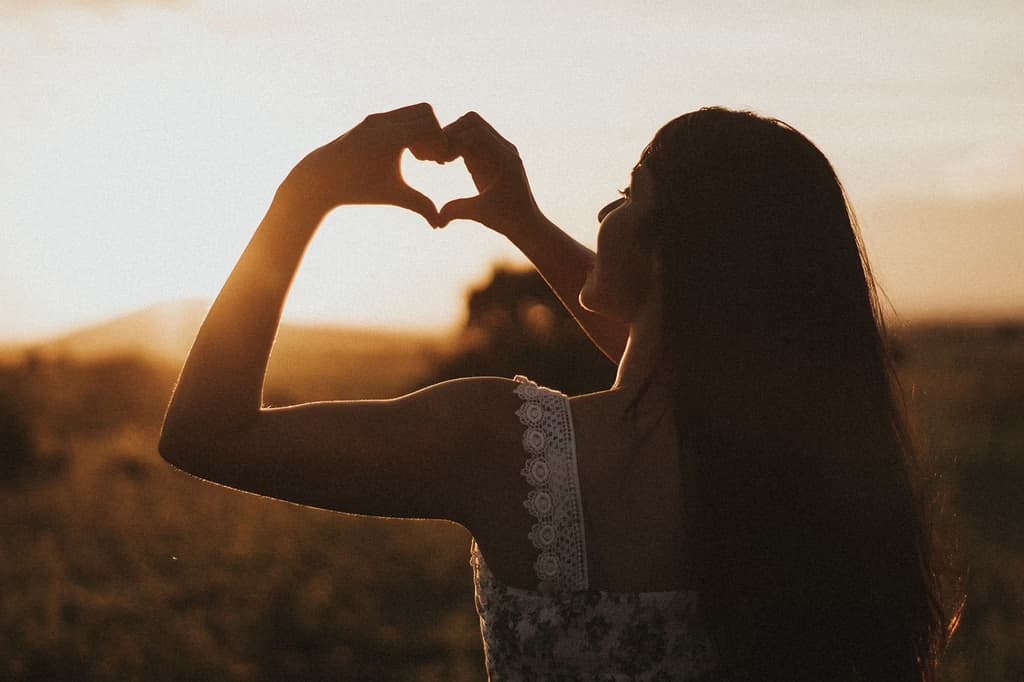 A woman stands outdoors at sunset, her long hair flowing. She forms a heart shape with her hands, silhouetted against the warm glow of the setting sun. The scene exudes a sense of peace and love.