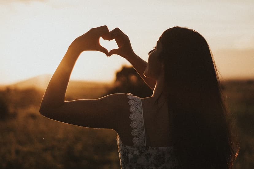 A woman stands outdoors at sunset, her long hair flowing. She forms a heart shape with her hands, silhouetted against the warm glow of the setting sun. The scene exudes a sense of peace and love.