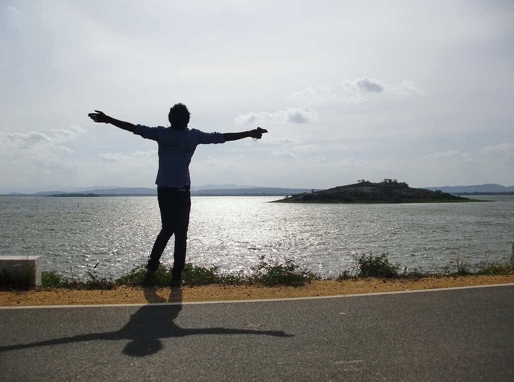 A person stands on a road near a body of water, arms outstretched, casting a shadow. The sun is low, causing the water to glisten. An island with trees is visible in the distance under a partly cloudy sky.