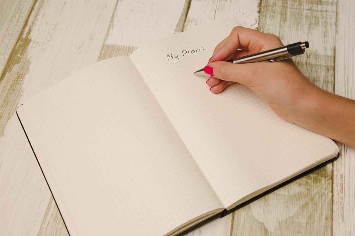 A hand with pink nail polish is writing My Plan: on the first page of a grid paper notebook with a pen. The notebook is open on a white wooden surface.