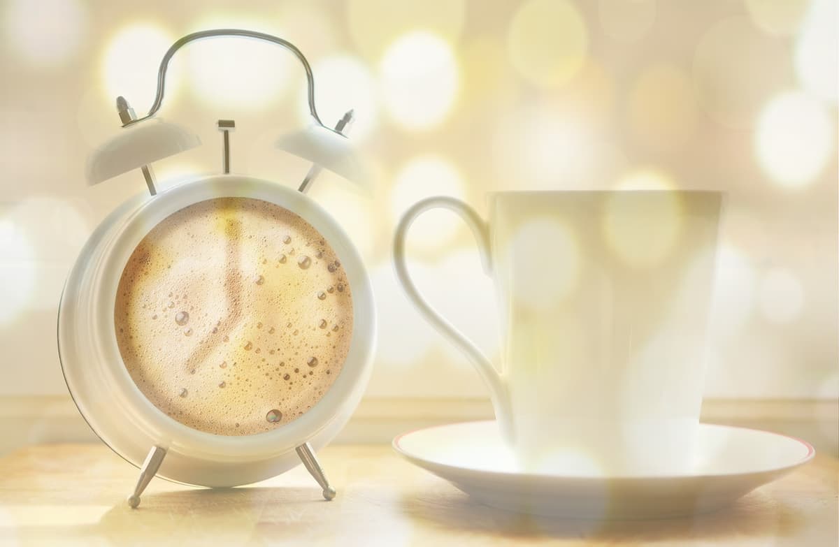 A white alarm clock with its face resembling a frothy coffee stands next to an empty white teacup on a saucer. The background is blurred with golden bokeh lights, creating a cozy, morning atmosphere.