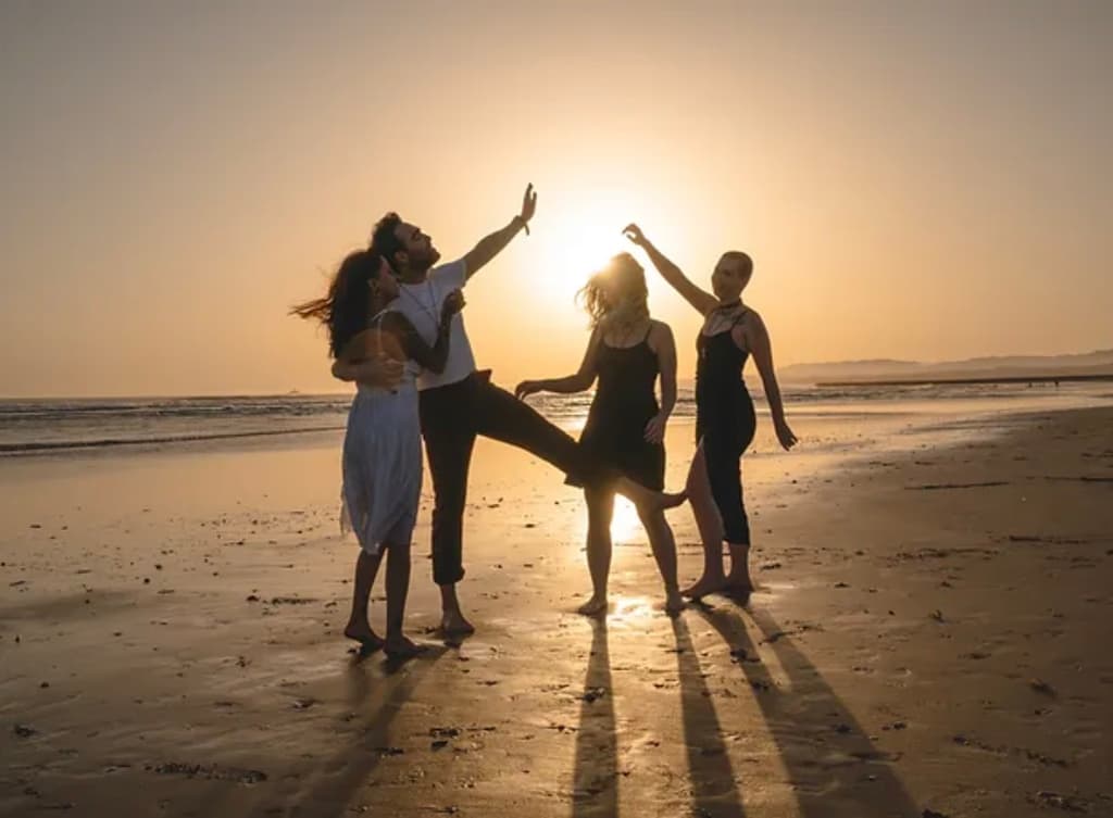 Four people joyfully dance in silhouette against a golden sunset on the beach, their movements casting long shadows on the sand. The sky is clear, and the sun glows brightly just above the horizon.