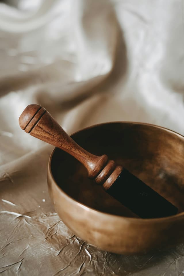A close-up of a brass singing bowl with a wooden mallet resting inside, placed on a textured, silky fabric surface in neutral tones.