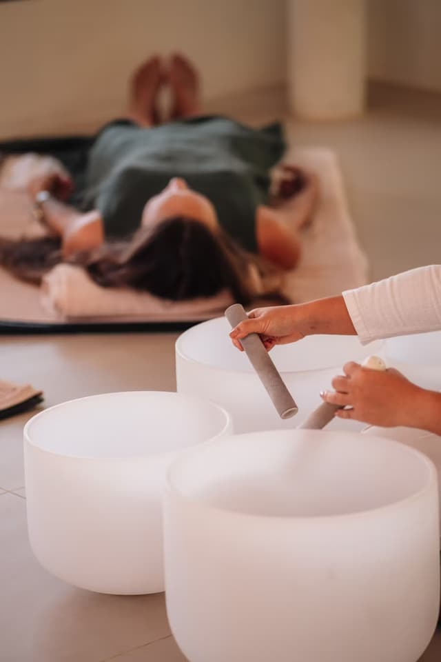 In a softly lit space, a sound therapist plays crystal singing bowls while a participant lies comfortably on a mat, enjoying a sound healing session.