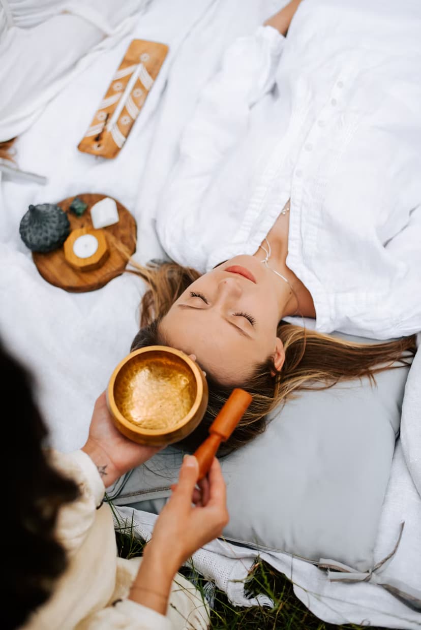 A person lying down on a soft surface with a singing bowl placed on their torso, surrounded by other singing bowls and a peaceful, softly lit environment, suggesting a sound healing or meditation session.