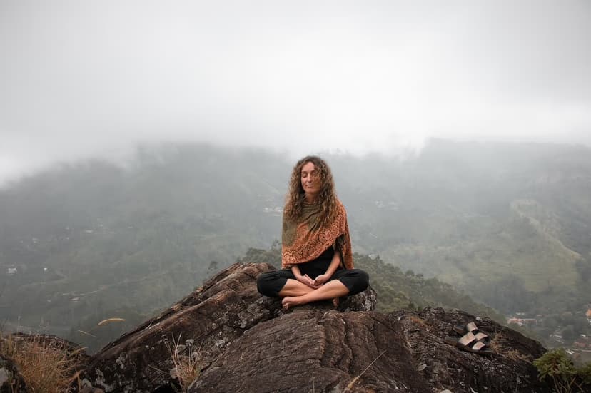 A person with curly hair sits cross-legged on a rock, meditating. They wear a shawl and dark pants. Misty hills and foggy skies fill the background, creating a serene atmosphere. Sandals lie beside them.